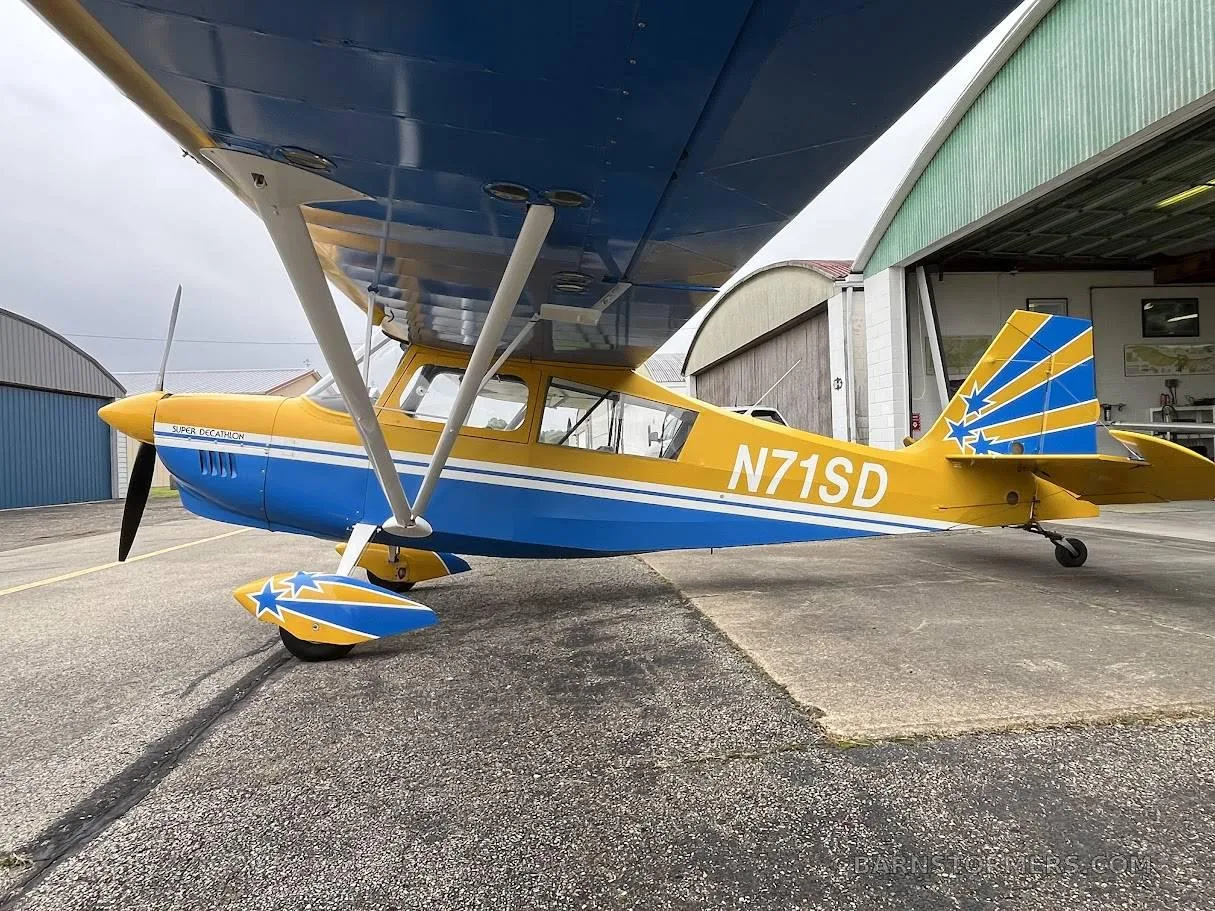 A yellow and blue Super Decathlon aircraft with tail number N71SD parked on an apron next to a hangar, viewed from the front-left.