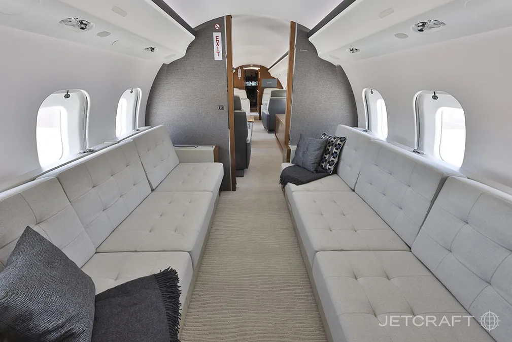 The spacious aircraft cabin features two long, light grey upholstered sofas with throw pillows facing each other.