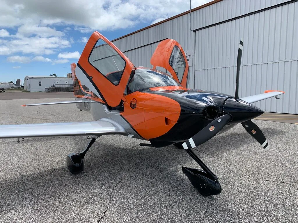 Front three-quarter view of a Cirrus SR20 aircraft with orange, black, and silver paint scheme, doors open, on an airfield.