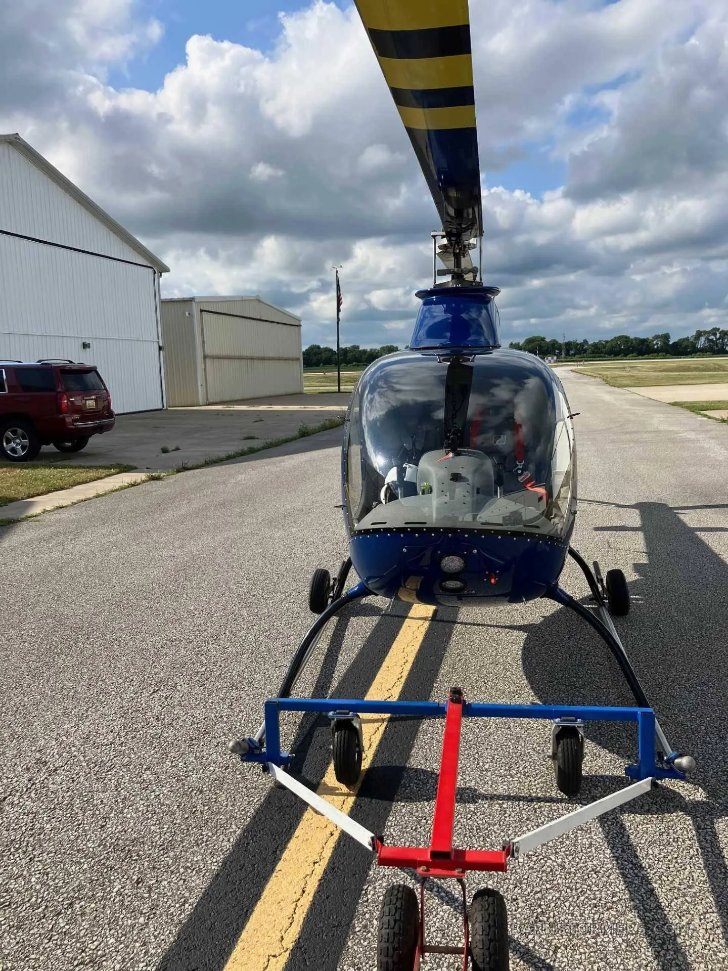 A blue helicopter (N911JE) with a yellow-striped rotor blade, viewed from the front, on an airfield tarmac with a ground handling cart.