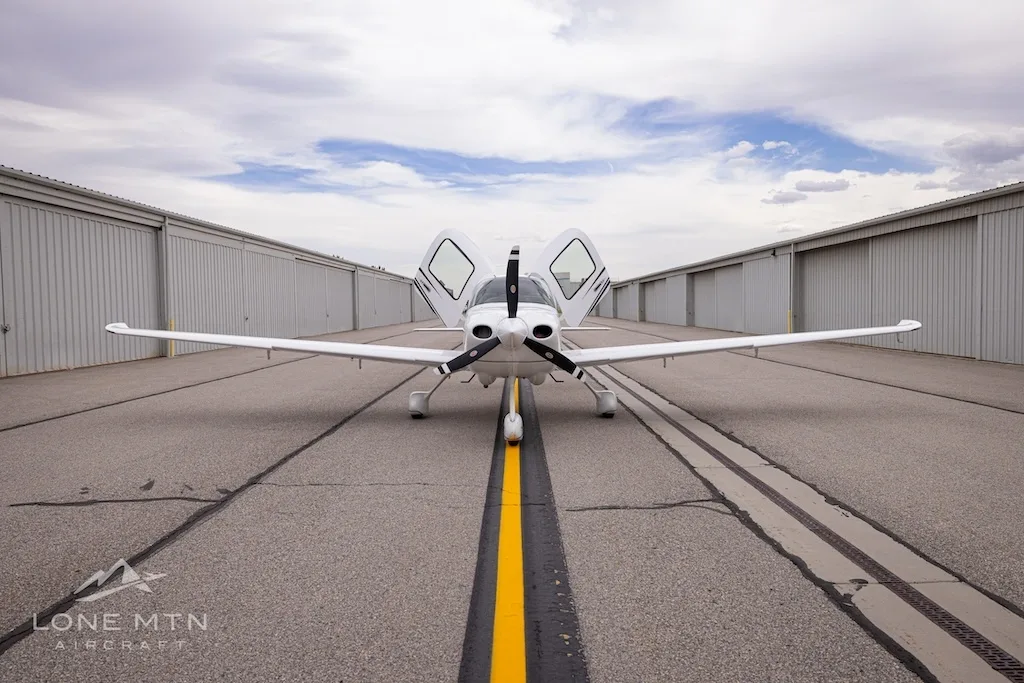 Head-on view of a white Cirrus SR22 aircraft with black and grey stripes, doors open, parked on an asphalt ramp.