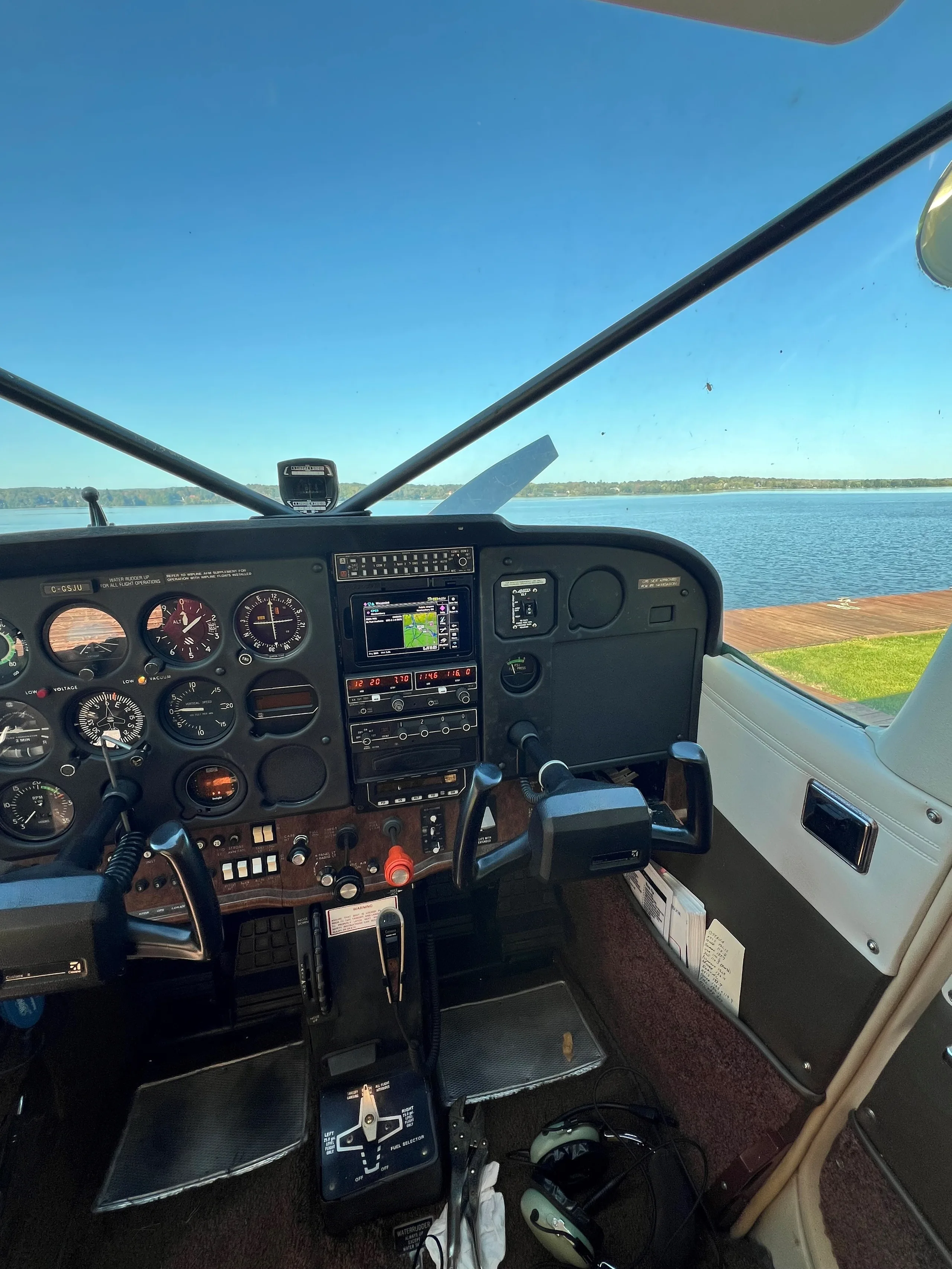 The cockpit of a floatplane shows the instrument panel and controls, with a view of a lake outside.
