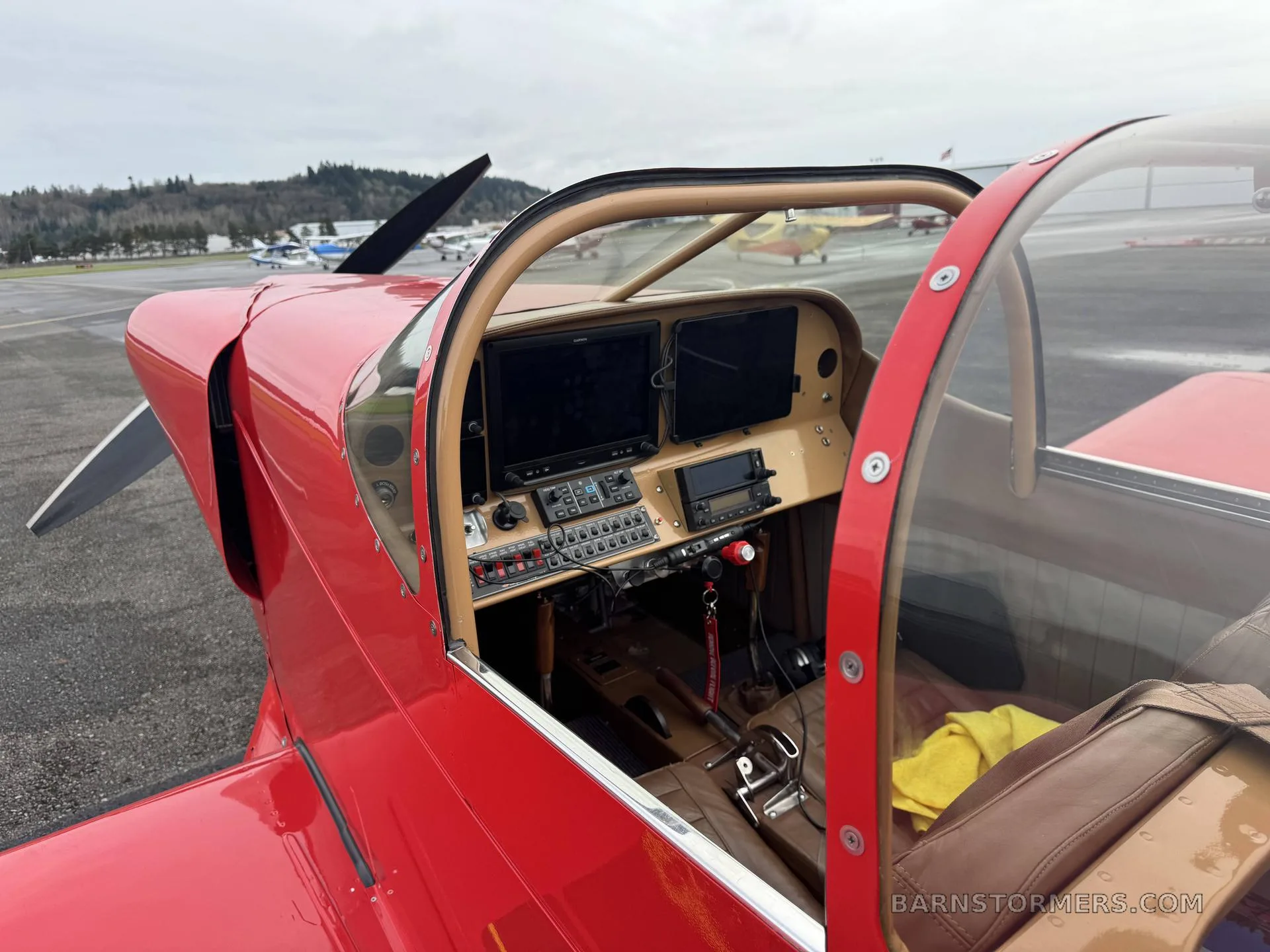 The cockpit of a red experimental aircraft features a modern glass panel with two large screens and various controls, seen through the open canopy.