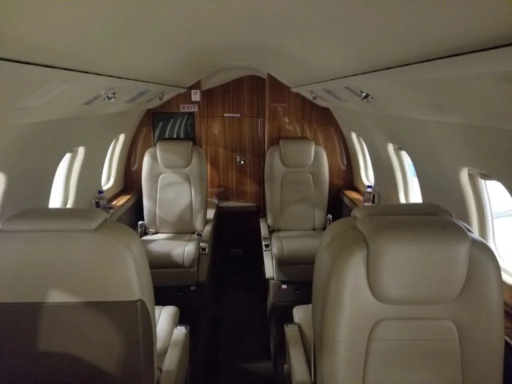 The interior cabin of a private jet with light beige leather seats and wood paneling, viewed from the front looking back.