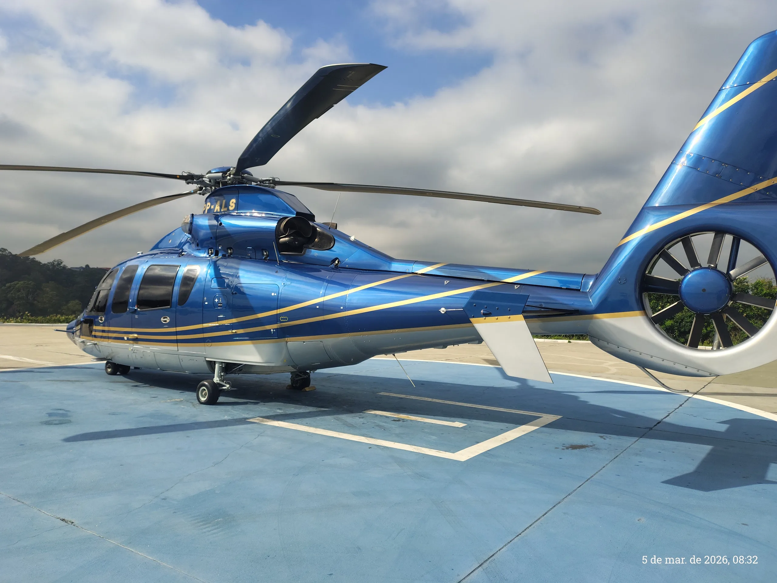A blue and silver helicopter with gold stripes is parked on a blue helipad under a cloudy sky, viewed from the rear-right.