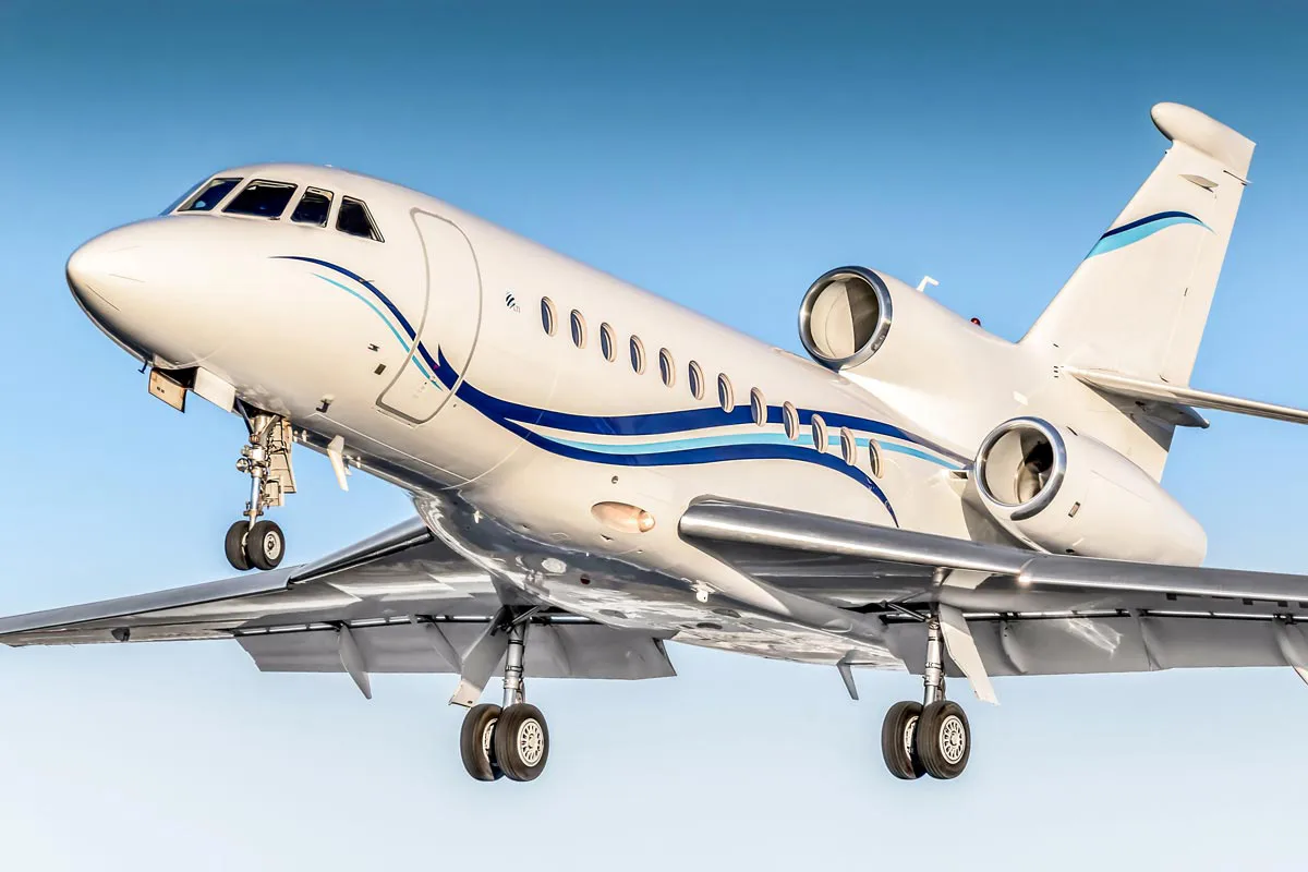 A white private jet with blue and silver stripes on its fuselage is captured in flight with its landing gear extended.