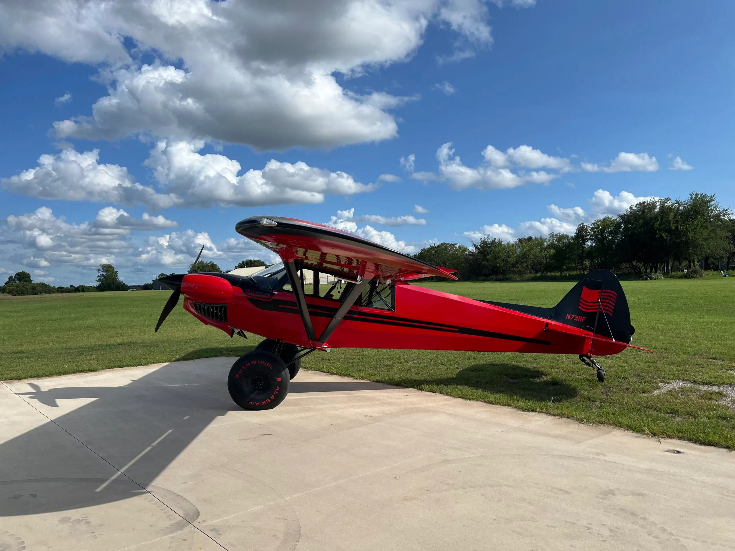 A red and black high-wing aircraft with large bush tires is parked on a concrete pad next to a grassy field under a partly cloudy sky.