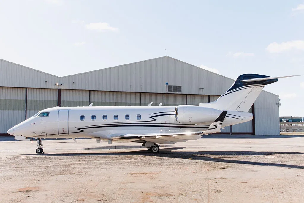 A white Bombardier Challenger 350 business jet with black and grey stripes parked on a sunny airfield in front of a hangar.