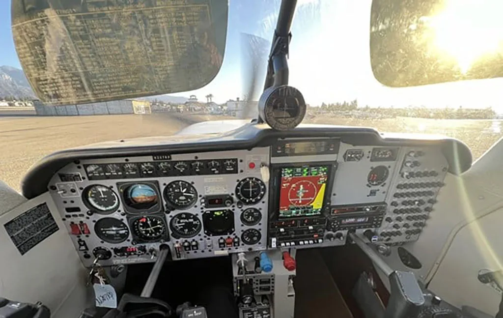 The cockpit of a single-engine aircraft shows a detailed instrument panel with both analog gauges and a modern digital display, with sunlight streaming through the windshield.