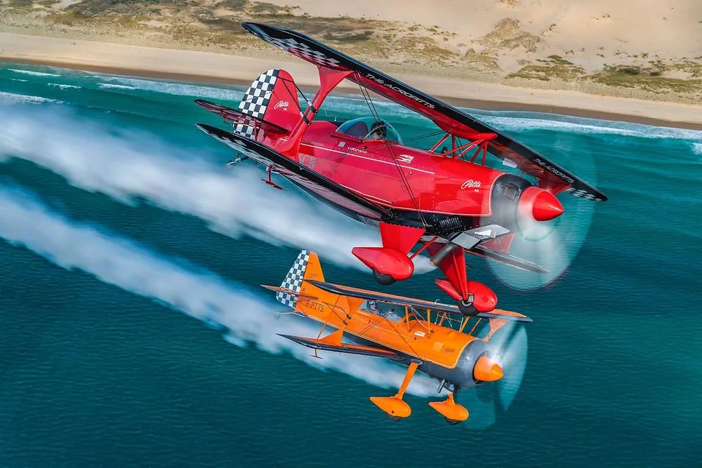 Two Pitts biplanes, one red and one orange, flying in formation over a coastline, leaving white smoke trails.
