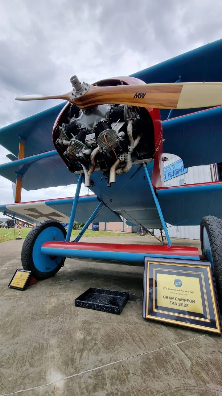 A close-up of the front of a red and blue Fokker Dr.I replica triplane, showing the exposed engine and propeller, with awards on the ground.