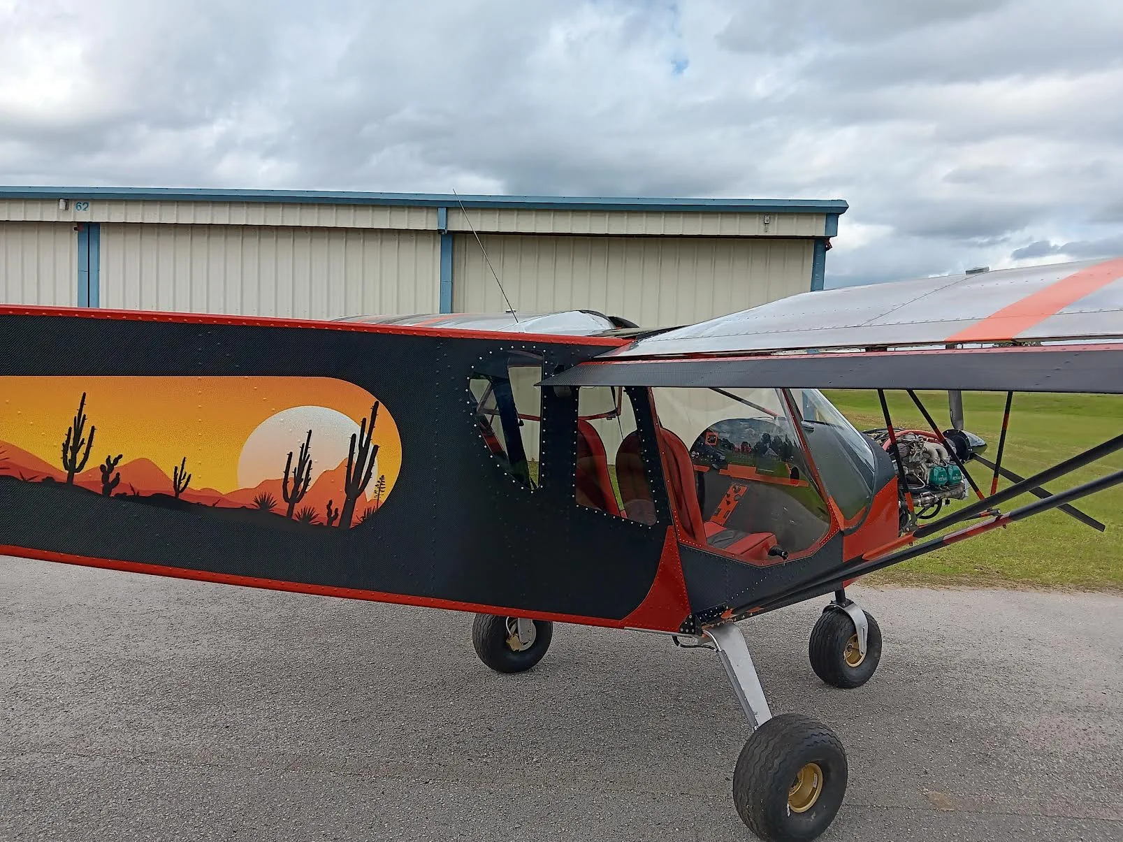 Side view of an orange and black ultralight aircraft with a desert sunset mural on its fuselage, parked next to a hangar.