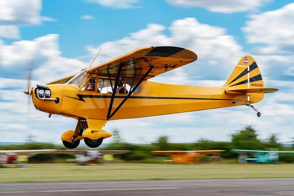 A yellow Piper Cub-style aircraft with black stripes, registration N30920, takes off from a grassy airfield under a blue sky with white clouds.