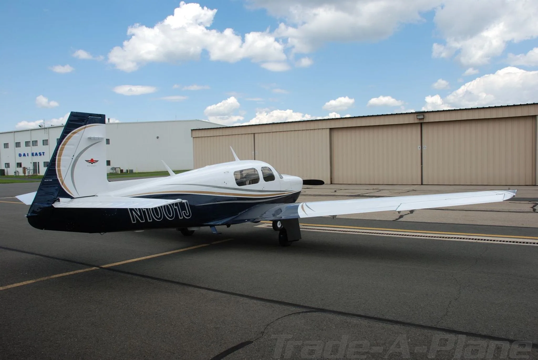 A white and blue Mooney aircraft (N1001J) seen from the rear, parked on a tarmac in front of hangars.
