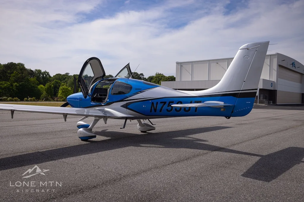 A blue and silver single-engine aircraft, N753JT, with open gull-wing doors is parked on an asphalt tarmac next to a hangar.