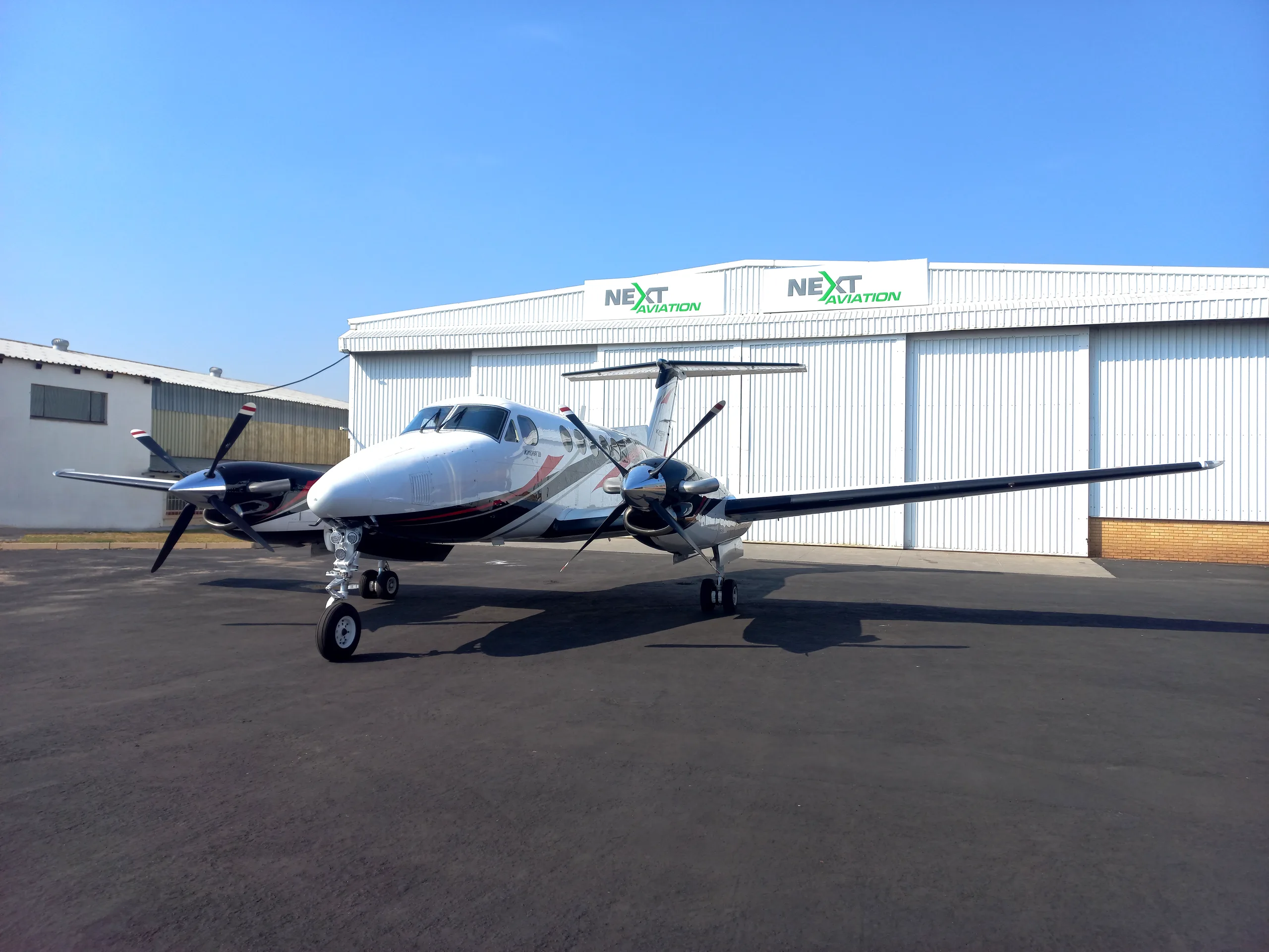 A white, black, and red Beechcraft King Air 200 twin-turboprop aircraft parked on an apron in front of a hangar.