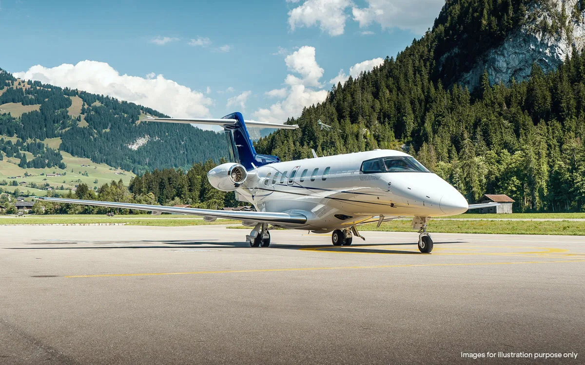 A white private jet with blue tail accents is parked on a tarmac with lush green mountains under a blue sky.