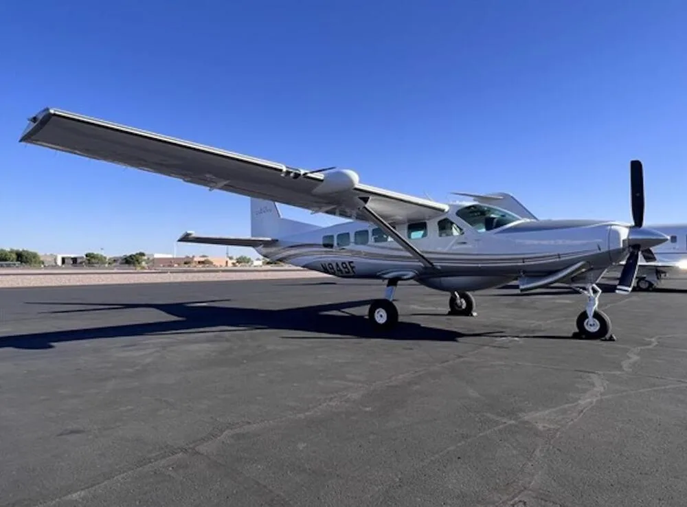 A silver Cessna Caravan N949F parked on an asphalt tarmac under a clear blue sky, viewed from the front-left.