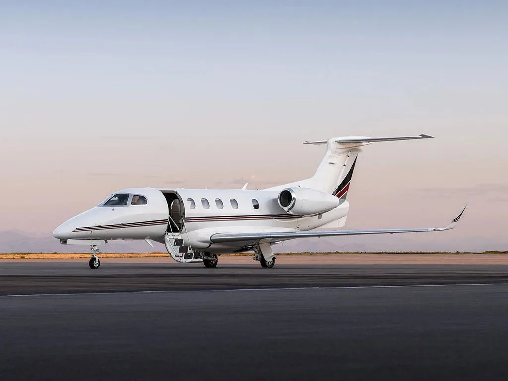 A white Embraer Phenom 300 business jet with red and black stripes, parked on an airfield with its main door open and stairs deployed.