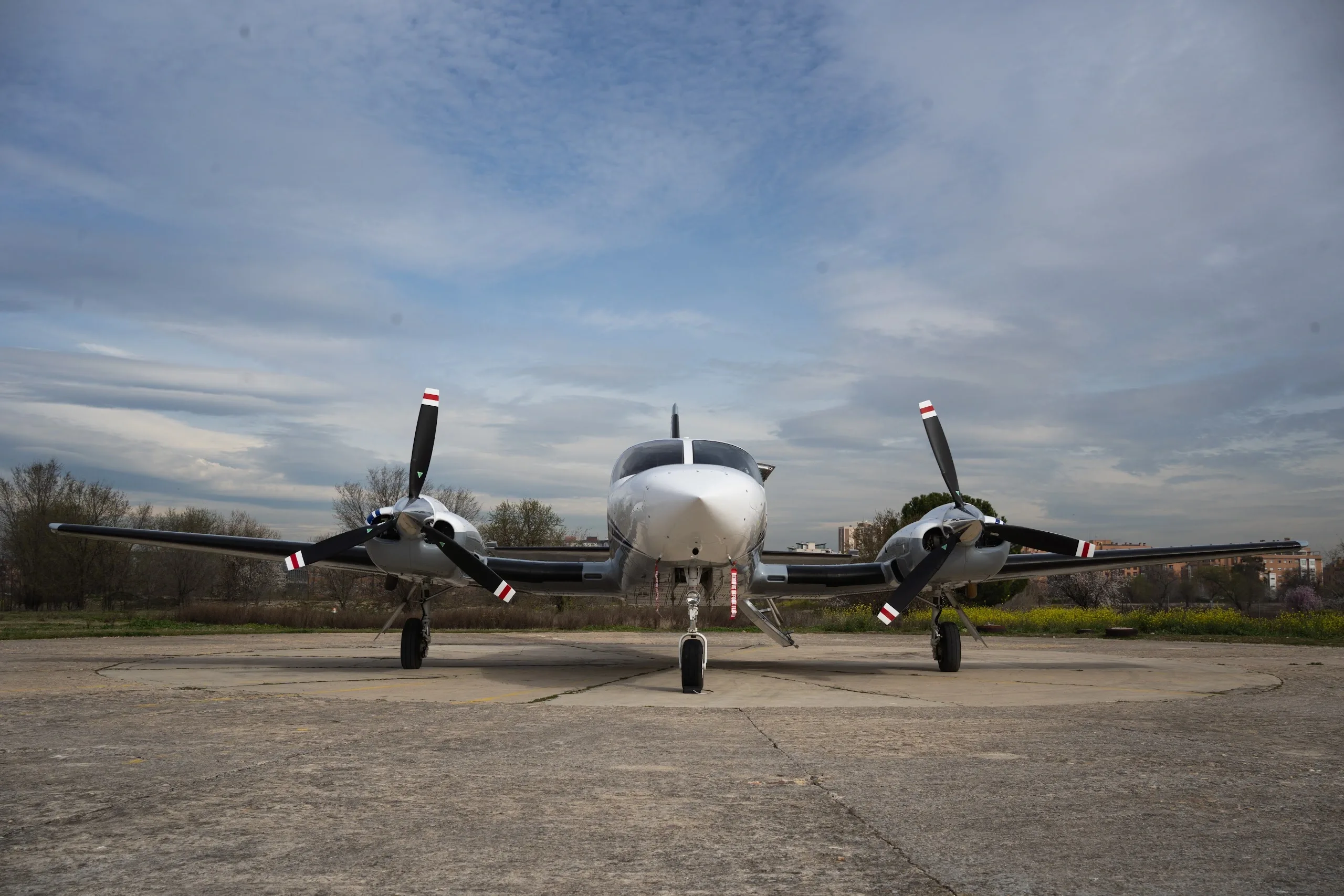 A front view of a white and grey twin-engine propeller aircraft with blue stripes parked on a concrete pad under a cloudy sky.