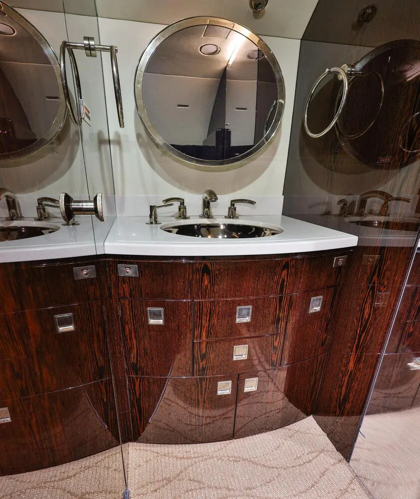 The aircraft lavatory featuring a white countertop with a sink, chrome fixtures, and dark wood cabinetry with mirrors.
