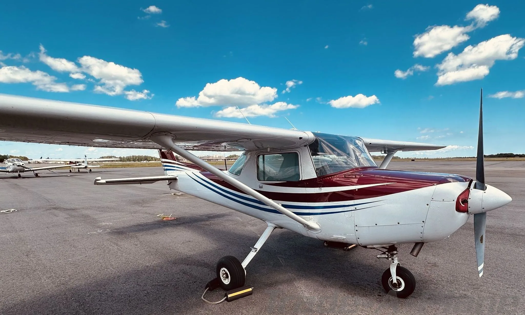 A white Cessna 172 with maroon and blue stripes on the fuselage and tail, parked on an airport tarmac under a blue sky.