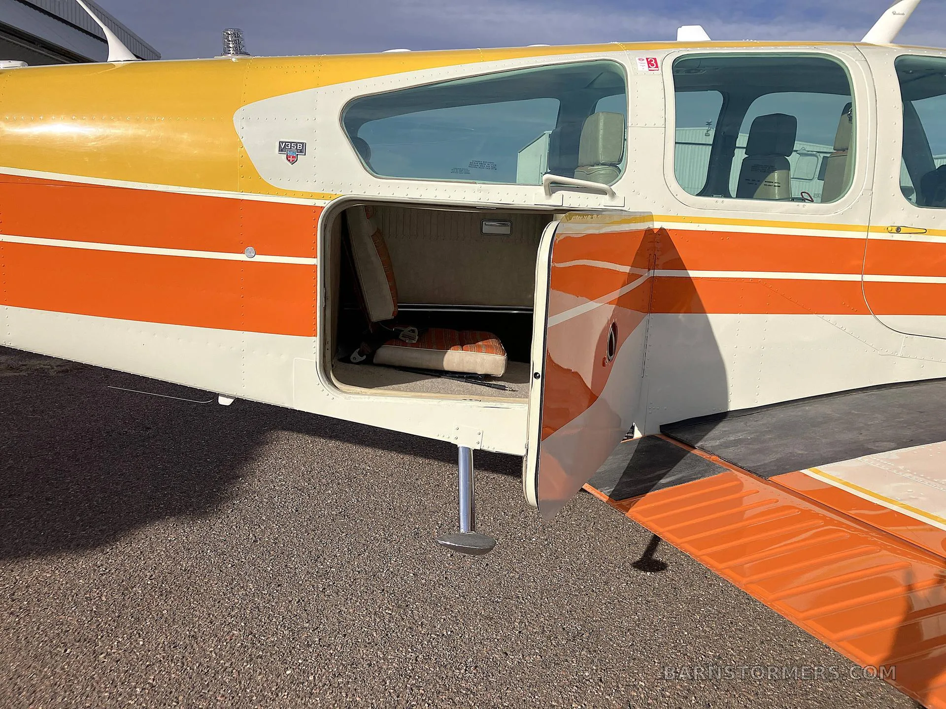 A close-up of the fuselage of a white, orange, and yellow Beechcraft Bonanza N725AP with its baggage compartment door open, revealing a small seat inside.