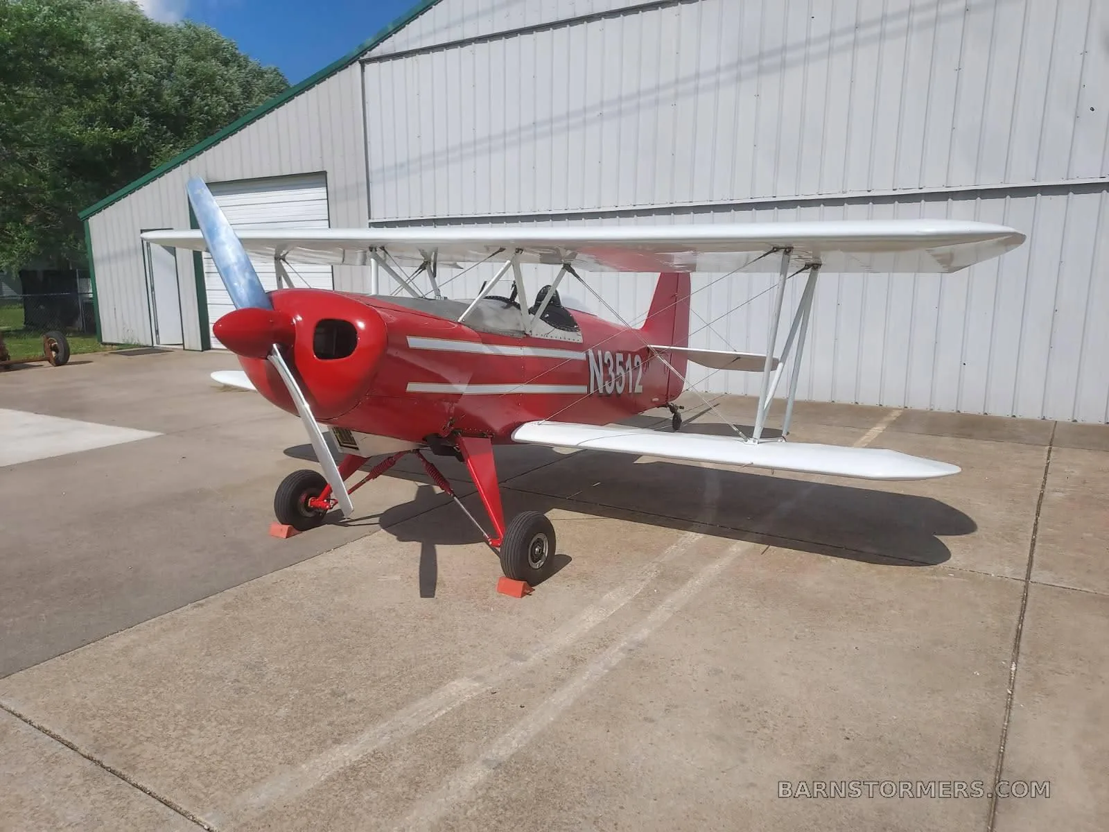 A red biplane with white wings and N3512 on the fuselage, parked on concrete next to a white hangar.