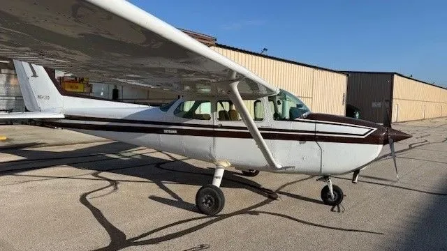 A white and brown Cessna Skyhawk (N64219) is parked on an airfield with hangars in the background under a clear sky.