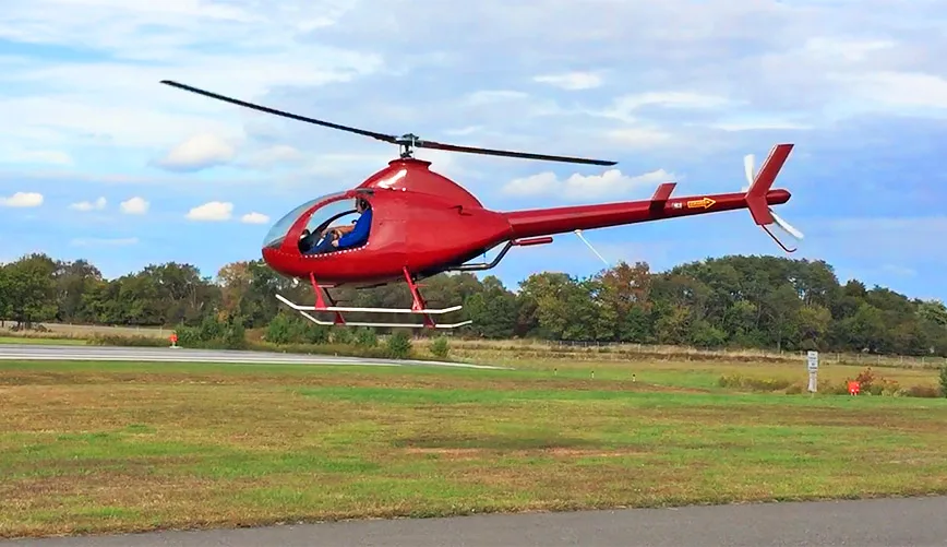 A red RotorWay Exec helicopter hovers over a grassy field near a runway.