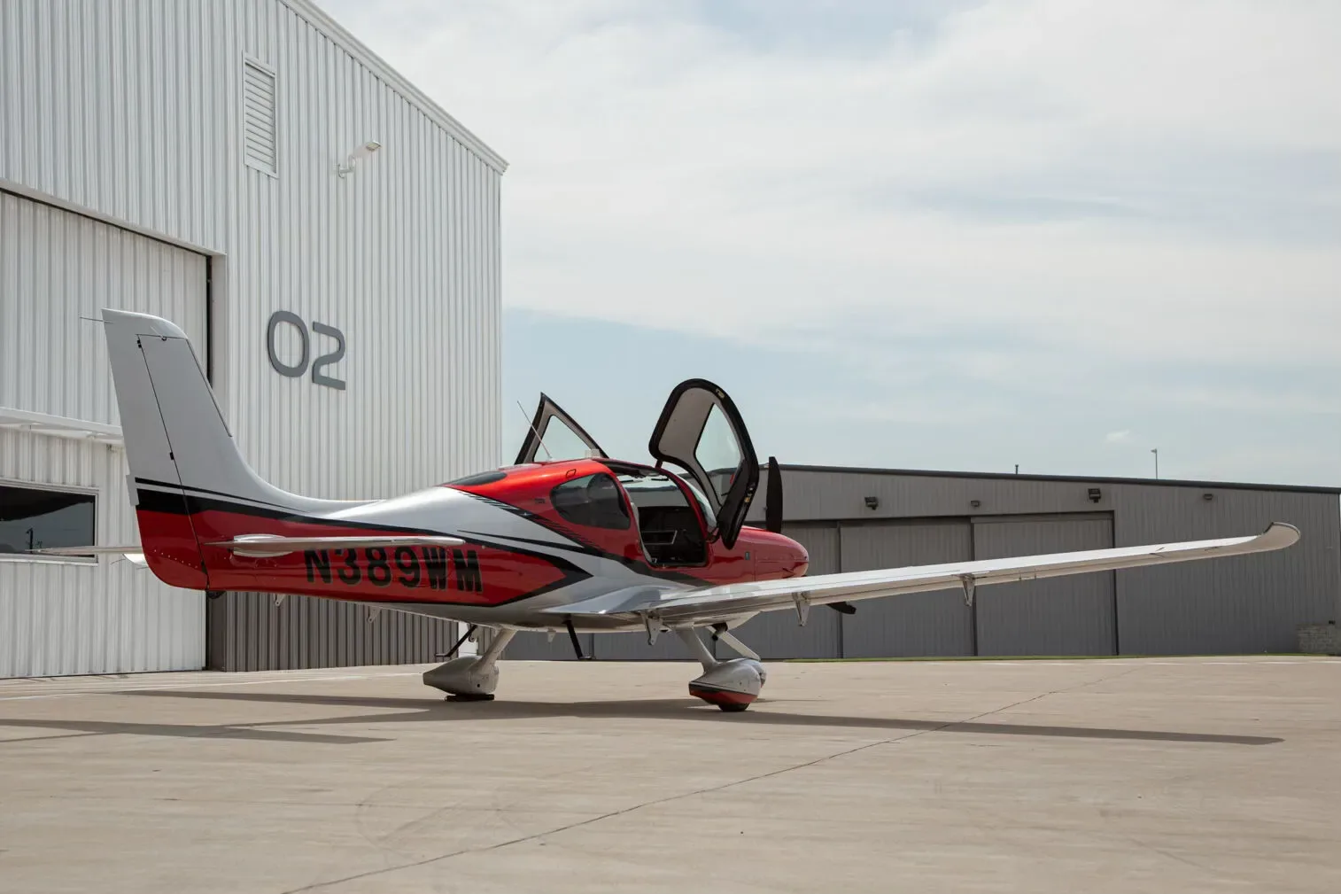 A red and silver single-engine propeller aircraft, N389WM, with its doors open, parked on an airfield next to a white hangar.