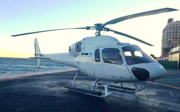 A white helicopter sits on a helipad with the ocean in the background.