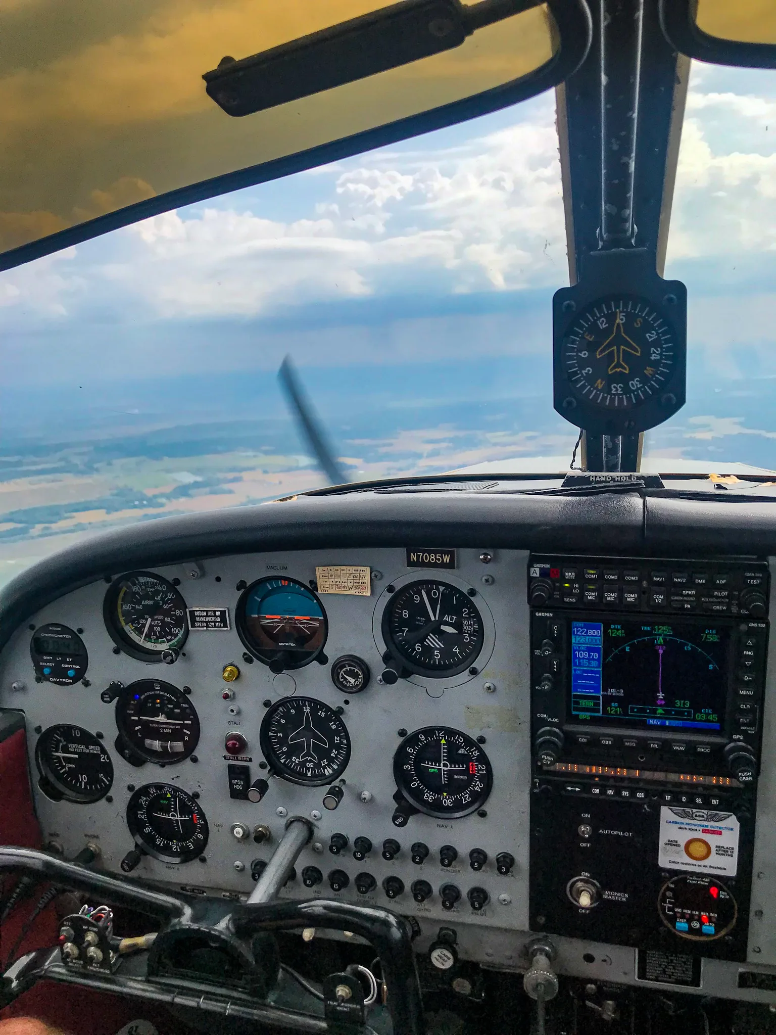 The cockpit instrument panel of a single-engine aircraft, showing gauges, radio stack, and a compass, with a view of clouds and land from the windshield.