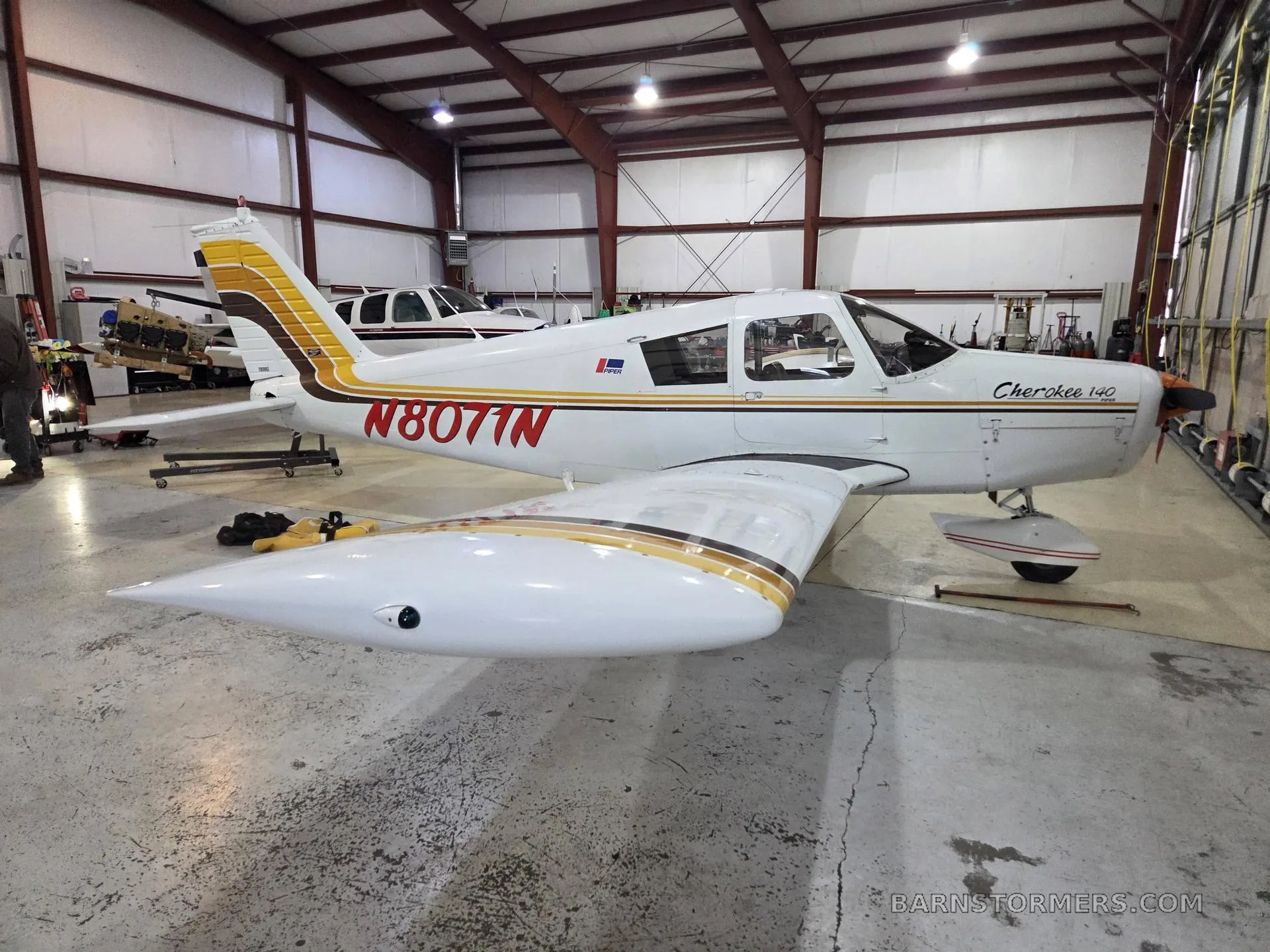 A white Piper Cherokee 140 with brown and gold stripes, registration N8071N, parked in a hangar.