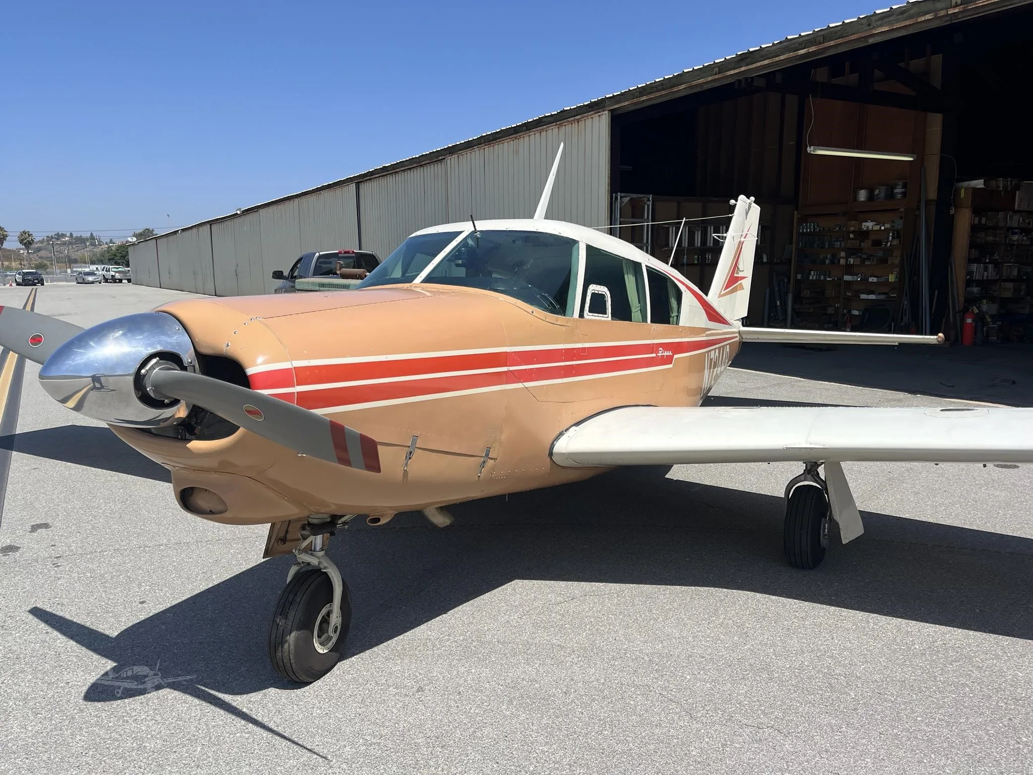 A tan and white single-engine Piper aircraft with red stripes is parked on an asphalt apron next to a hangar on a sunny day.