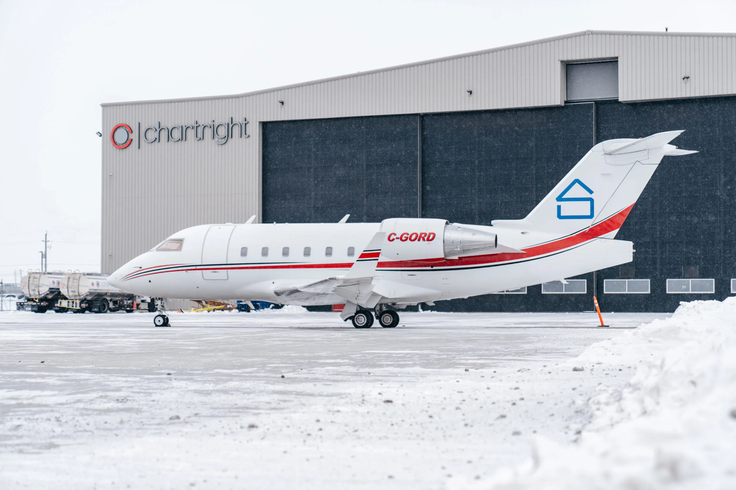 A white business jet, registration C-GORD, with red and black stripes and a blue house logo on the tail, is parked on a snowy tarmac in front of a hangar.
