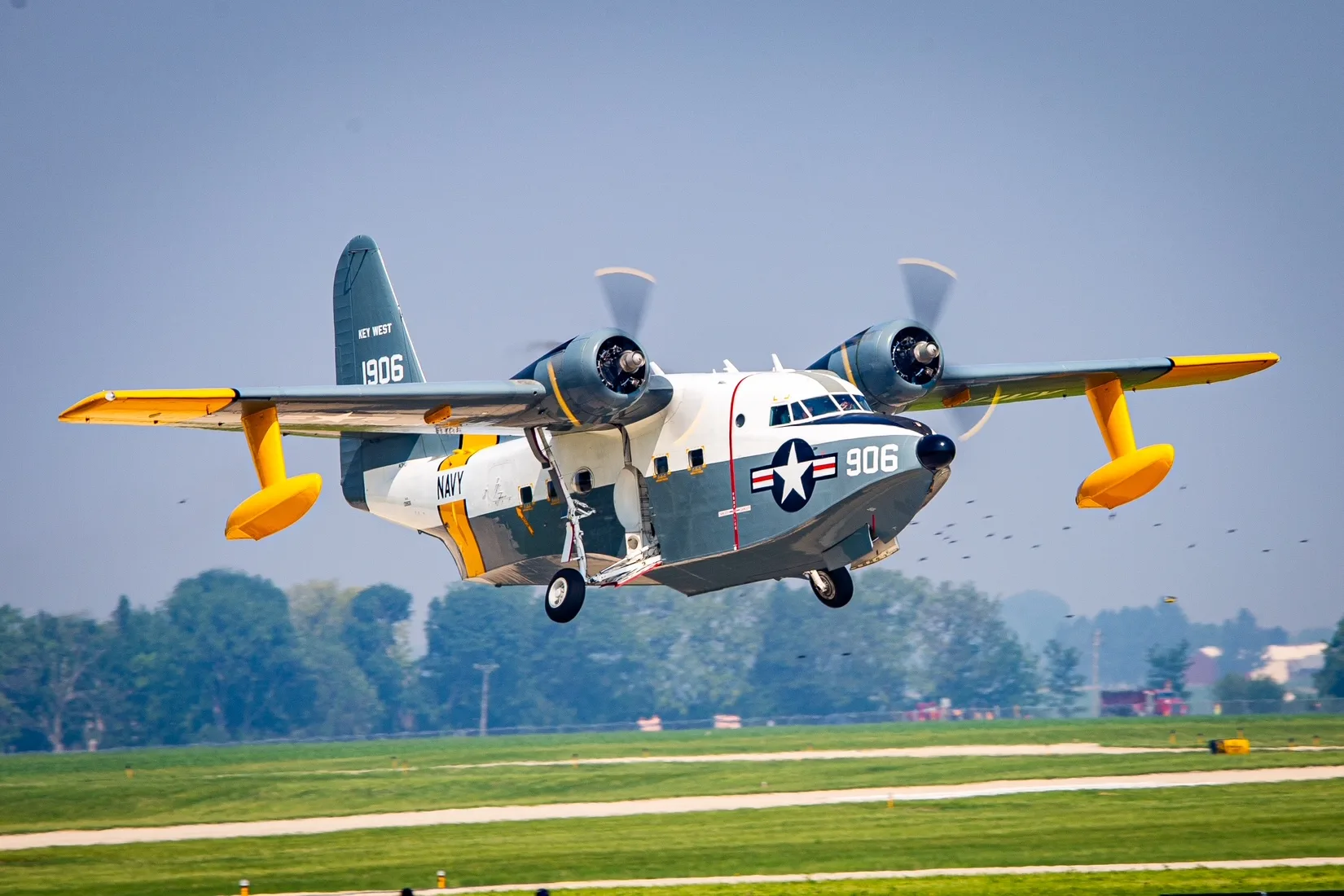 A Grumman HU-16 Albatross seaplane in US Navy livery with '906' on the nose, flying low over a green airfield with landing gear extended.