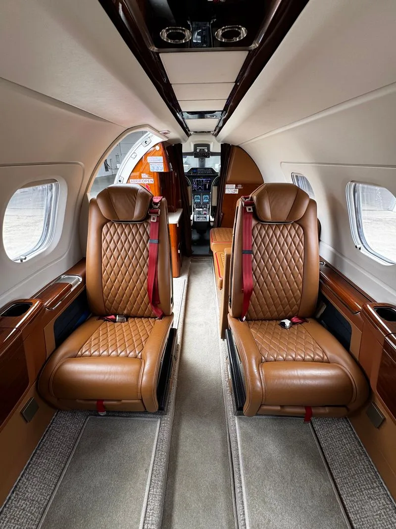 The aircraft cabin features quilted brown leather seats with red seatbelts, looking forward towards the open cockpit door.