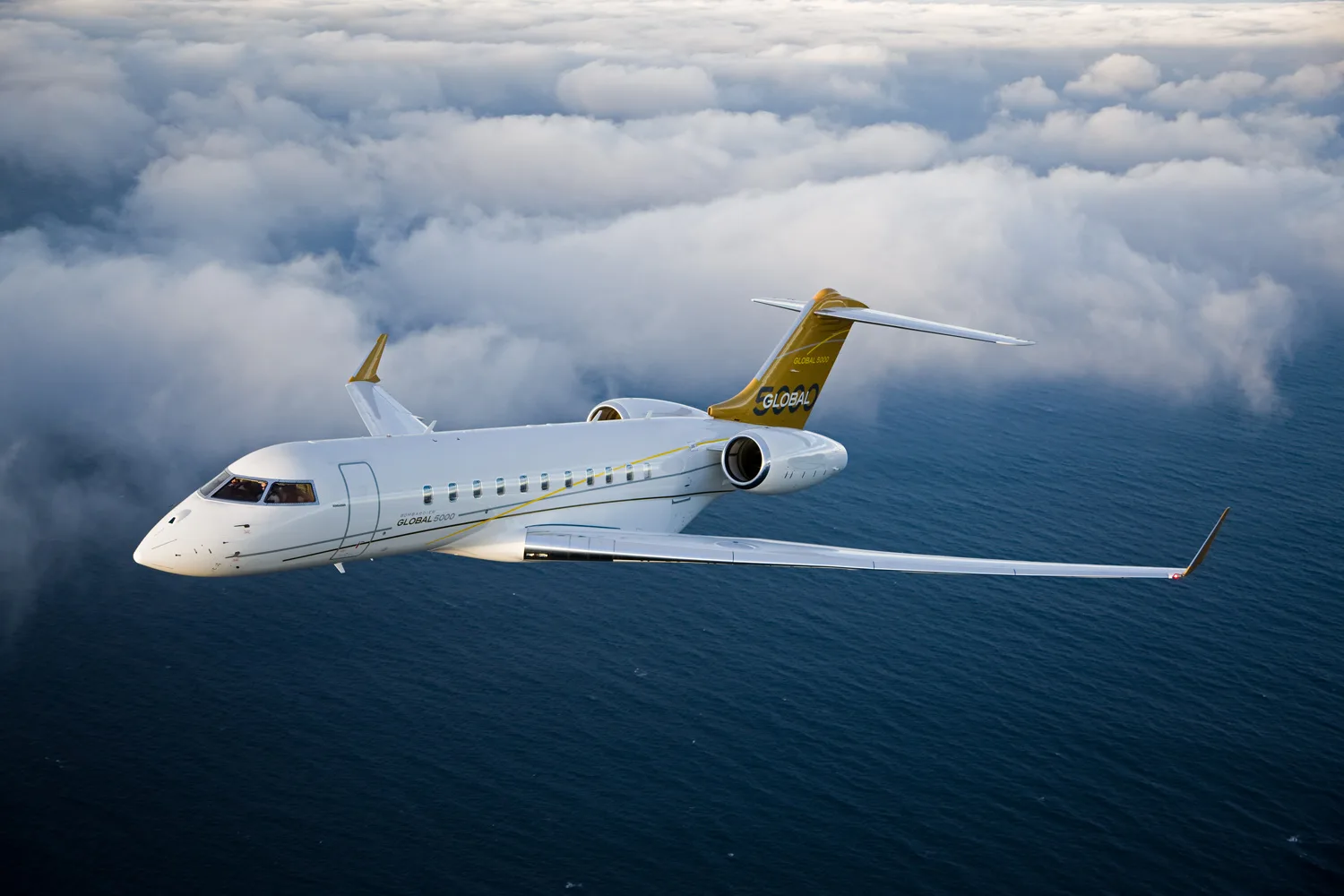 A white Bombardier Global 5000 business jet with gold tail accents flies above the ocean with clouds in the background.