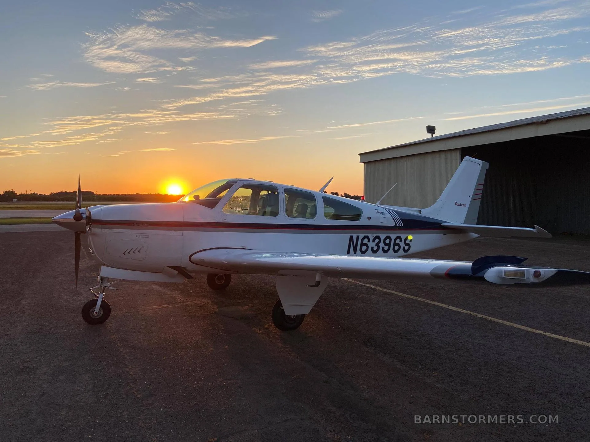 A white and blue Beechcraft Debonair N6396S parked on an tarmac at sunset, with a hangar in the background.