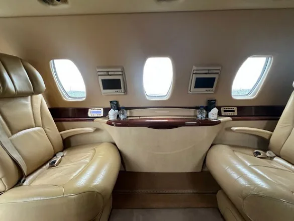 The aircraft cabin features two tan leather seats facing each other with a wooden table in between and small monitors above the windows.