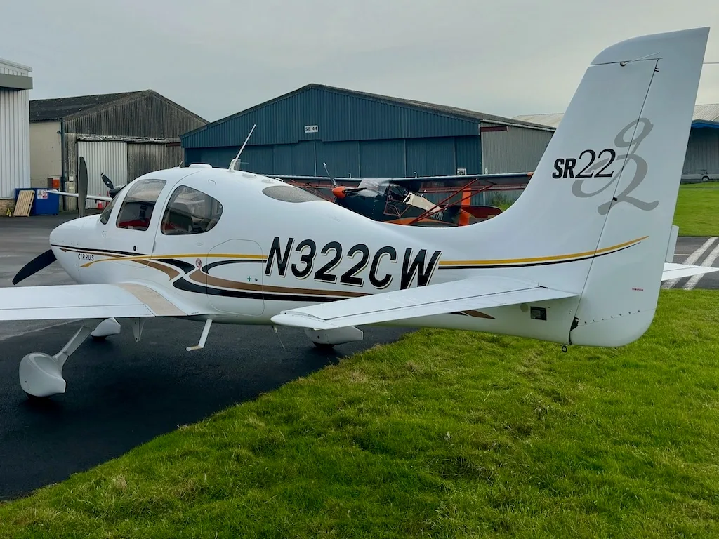 A rear-side view of a white Cirrus SR22 aircraft with N322CW registration, parked on a tarmac next to green grass.