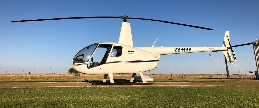 A white Robinson R44 helicopter with registration ZS-HYR parked on a paved area in a grassy, open field.