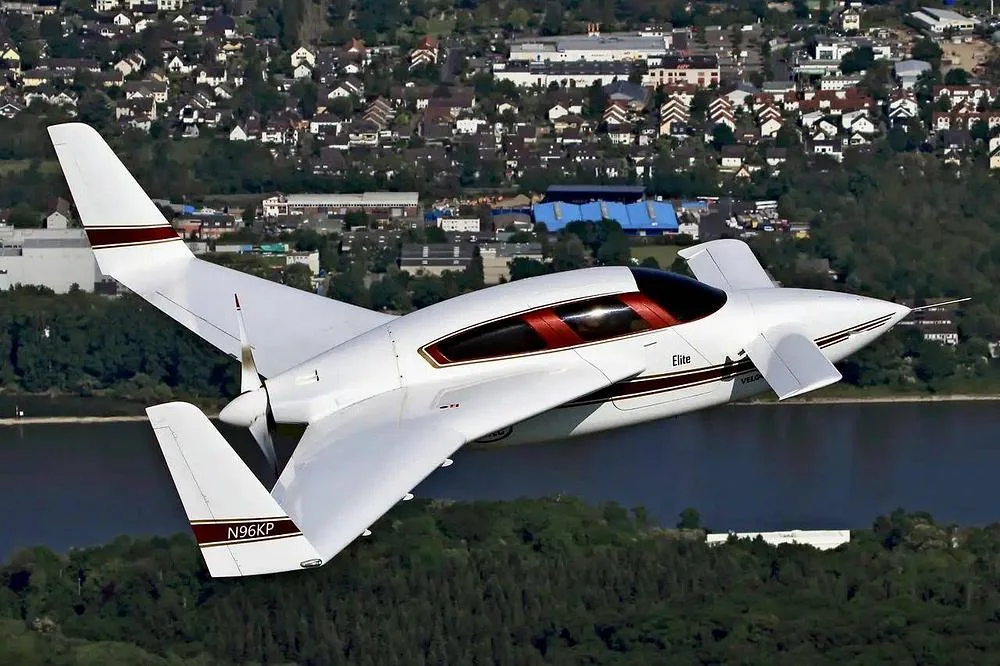 A white Velocity Elite aircraft with red and gold stripes, registration N96KP, flying over a city and water.