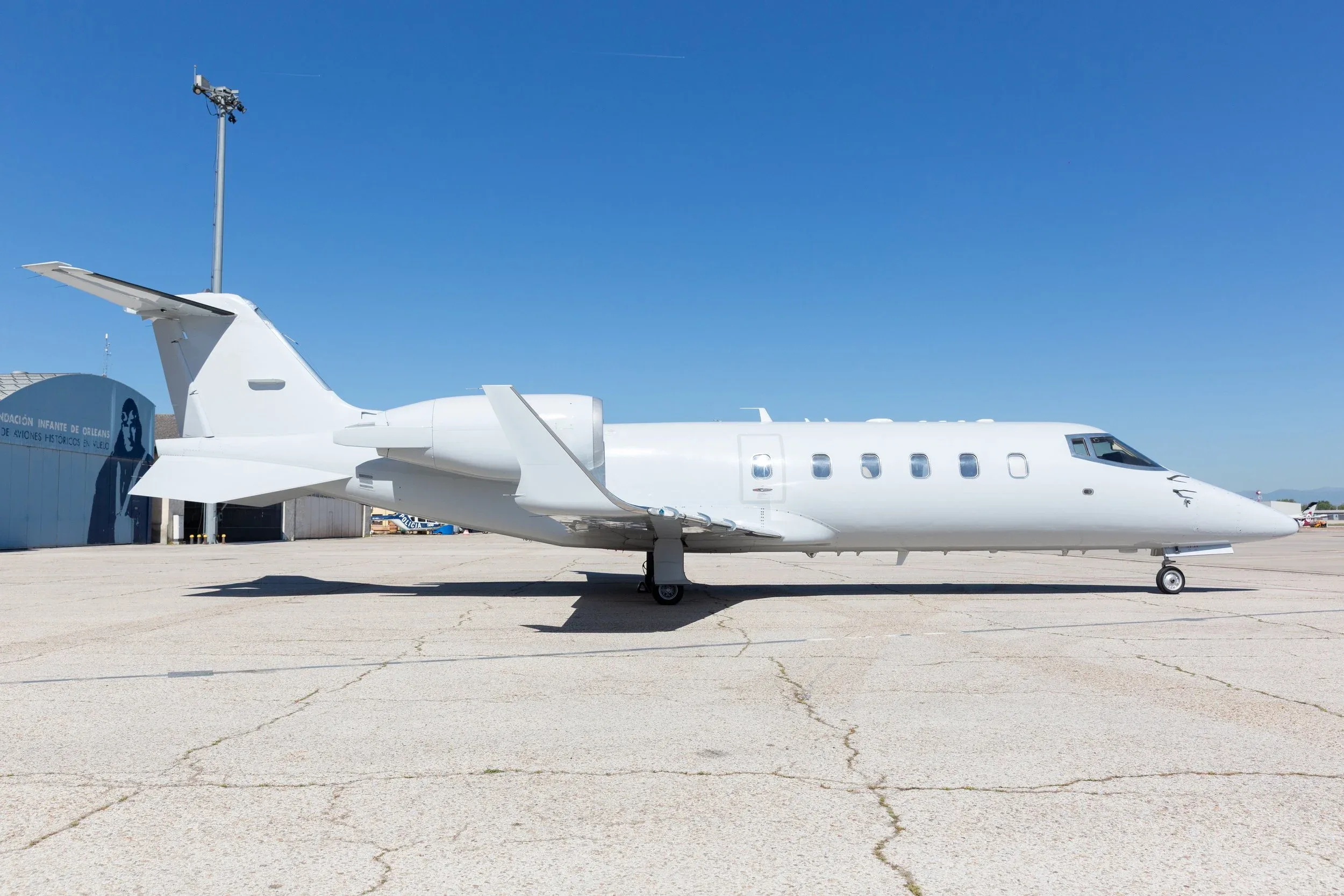 A white business jet is parked on a tarmac with a hangar and clear blue sky in the background.