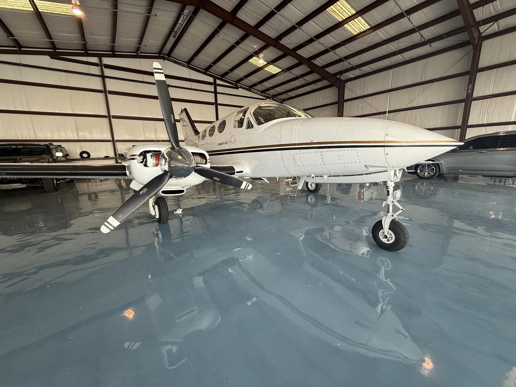 A white twin-engine turboprop aircraft with gold stripes is parked inside a hangar with a shiny gray floor.