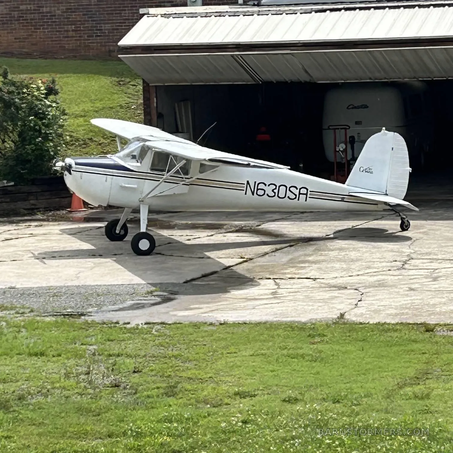 A white Cessna 170B with dark blue and gold stripes is parked on a concrete pad outside a garage.
