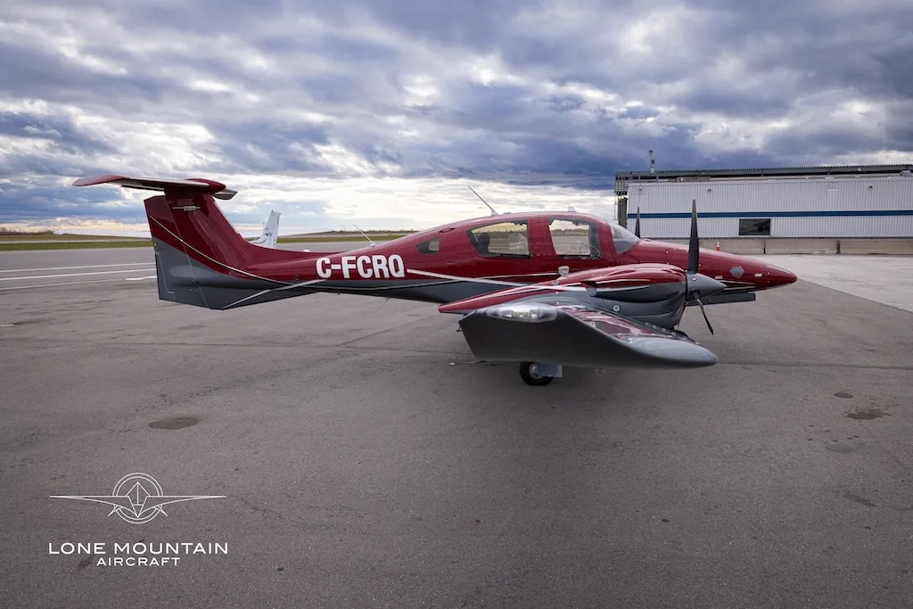 A red and grey Diamond DA62 aircraft with registration C-FCRQ parked on an airfield next to a hangar.