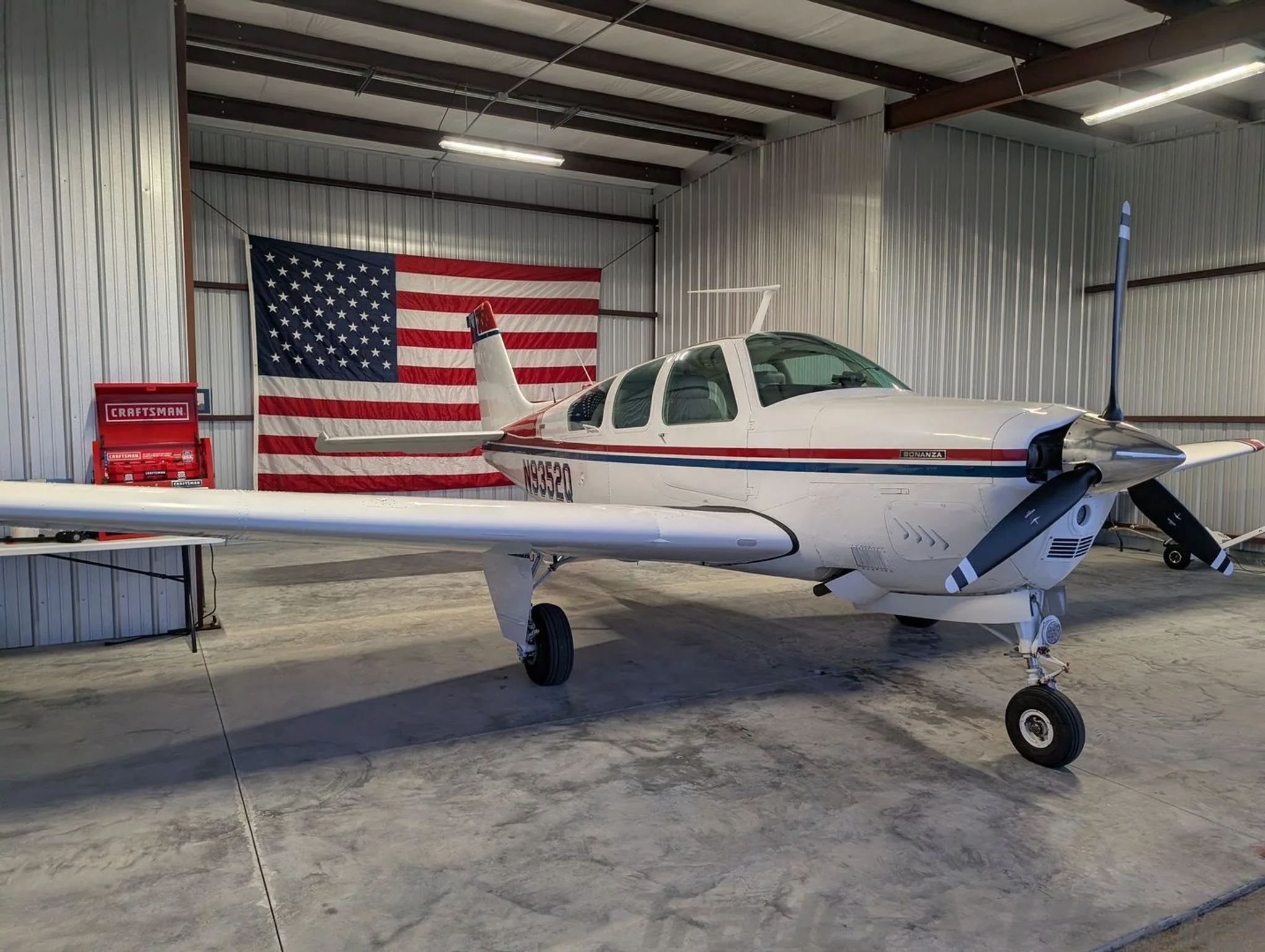 The aircraft is parked in a hangar with an American flag in the background.