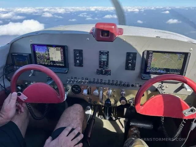 View from the cockpit showing the instrument panel with two red yokes and digital displays, flying above clouds.